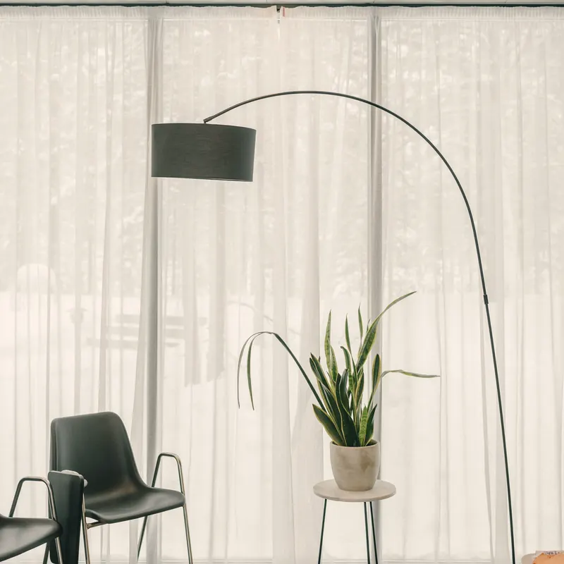 Sala de espera clara com luminária em arco, poltrona, planta e cortina translúcida filtrando a luz.